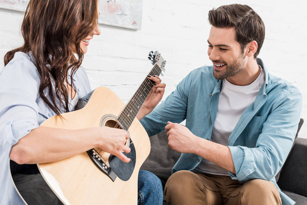 happy couple sitting on couch and playing acoustic guitar at home - Photo, Image