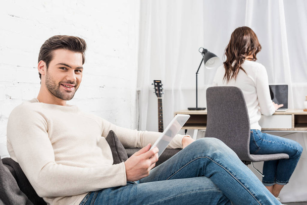smiling man using digital tablet while woman sitting at computer desk on background in living room - Foto, Imagem