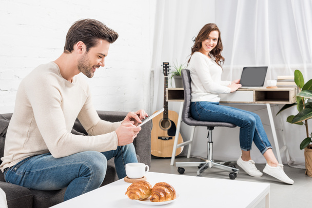 Mann nutzt digitales Tablet, während schöne Frau am Computer-Schreibtisch im Wohnzimmer sitzt - Foto, Bild
