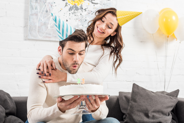 背景にパーティー ハットで笑顔の女性の誕生日ケーキのろうそくを吹きの男 - 写真・画像