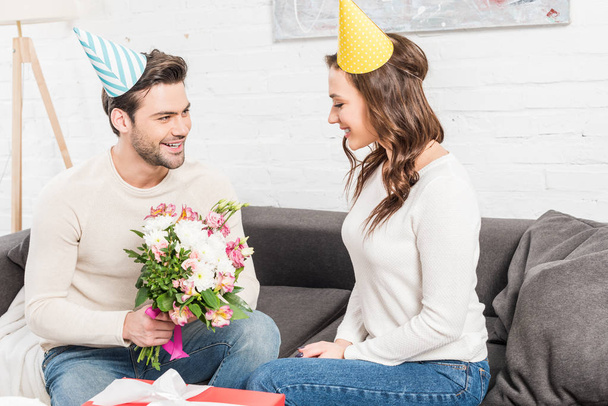 handsome smiling man in party hat presenting birthday flowers to woman in living room - Photo, Image
