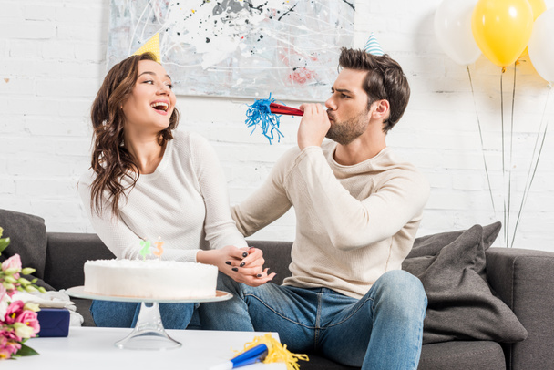 couple in party hats celebrating birthday with cake while man blowing party horn at home - Photo, Image
