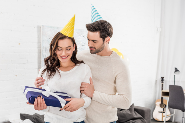 smiling couple in party hats embracing while holding birthday present in living room - Fotografie, Obrázek