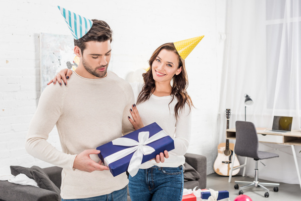 beautiful couple with gift box embracing and celebrating birthday in living room - Photo, Image