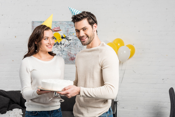 beautiful couple in party hats holding cake and celebrating birthday in living room - Fotó, kép