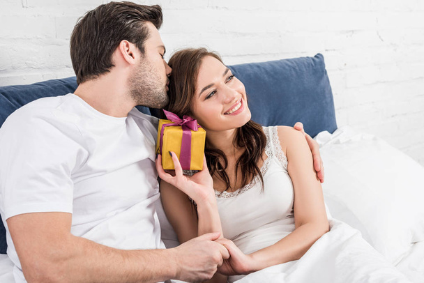 couple in nightwear lying in bed, hugging and holding birthday present at home  - Фото, изображение