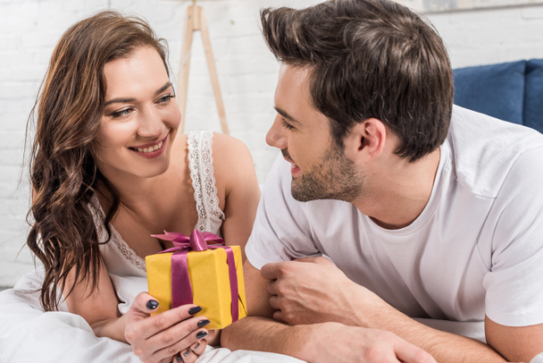 happy couple lying in bed, holding birthday present and looking at each other at home  - Foto, Bild
