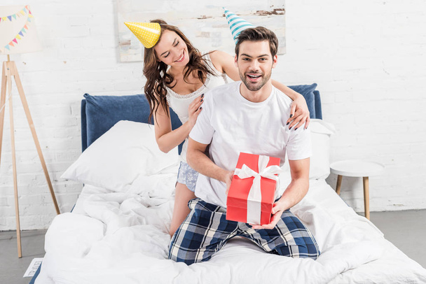happy couple in party hats celebrating event and holding birthday present in bed - Foto, afbeelding