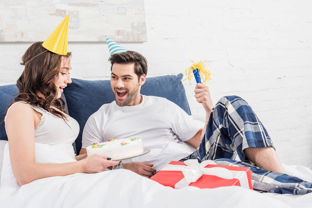 excited couple in party hats lying in bed and celebrating birthday with cake and party horn at home - Photo, Image