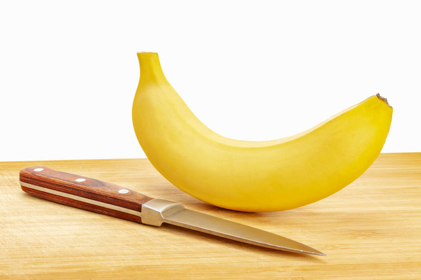 A fresh banana with a knife is lying on a wooden board. The background is white, isolate - Фото, зображення