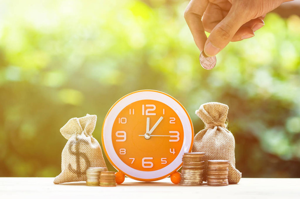 Geld sparen, investieren, Zeit und Geld wachsen Konzept: Stapeln wachsenden Münzen, Geldbeutel und orangefarbene Uhr auf Holztisch. spart Geld für die Zukunft. Zeitinvestitionen verwenden Geld für Arbeit. - Foto, Bild