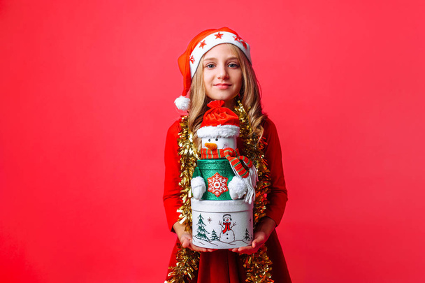 Teenage girl wearing Santa hat and tinsel on neck holding Christmas gift boxes, on red background - Valokuva, kuva