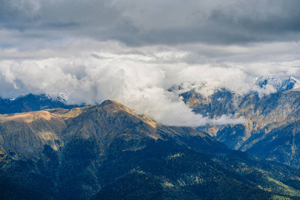 Mountain landscape. Warm autumn Sunny day. The slopes of the mountains covered with autumn woods. Clouds above mountain peaks. The snow on the tops - Photo, image