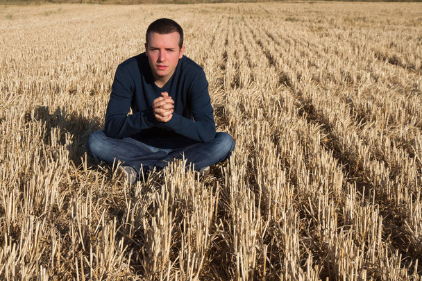 Joven sentado en el suelo en el campo de cereales segados con los dedos entrelazados de las manos y mirando a la cámara
 - Foto, imagen