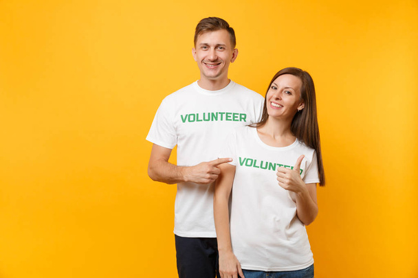 Portrait of young colleagues couple in white t-shirt with written inscription green title volunteer isolated on yellow background. Voluntary free work, assistance help, charity grace teamwork concept - Foto, imagen