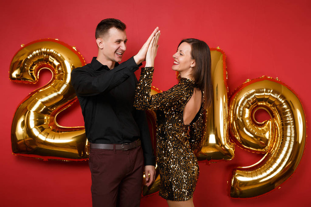 Couple guy girl in shiny glitter dress, black shirt celebrating holiday party isolated on bright red wall background golden numbers air balloons studio portrait. Happy New Year 2019 Christmas concept - Photo, Image