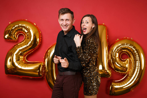 Couple guy girl in shiny glitter dress, black shirt celebrating holiday party isolated on bright red wall background golden numbers air balloons studio portrait. Happy New Year 2019 Christmas concept - Valokuva, kuva