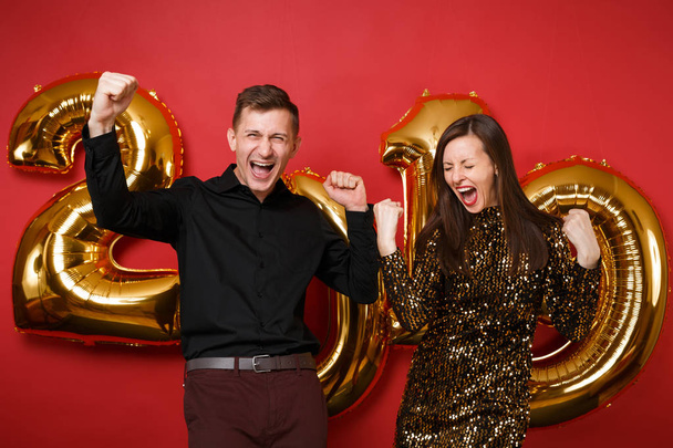 Couple guy girl in shiny glitter dress, black shirt celebrating holiday party isolated on bright red wall background golden numbers air balloons studio portrait. Happy New Year 2019 Christmas concept - Photo, Image