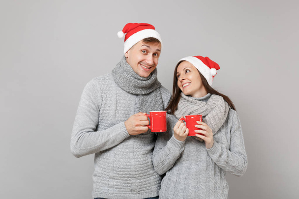 Fun couple girl guy in red Santa Christmas hat gray sweaters scarves hold cups of tea isolated on grey wall background, studio portrait. Happy New Year 2019 holiday party concept. Mock up copy space - Фото, изображение