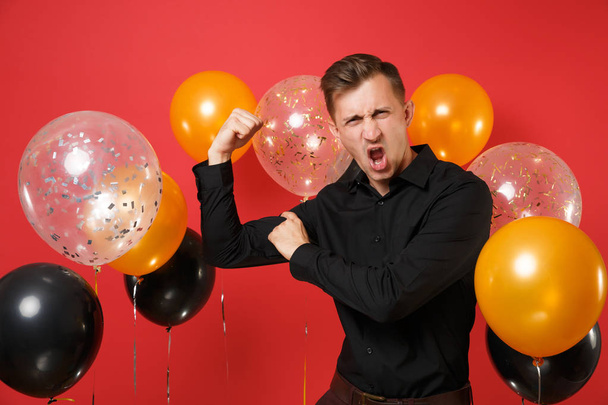 Strong young man in black classic shirt showing and pointing index finger on biceps on bright red background air balloons. St. Valentine's Day, Happy New Year birthday mockup holiday party concept - Photo, Image