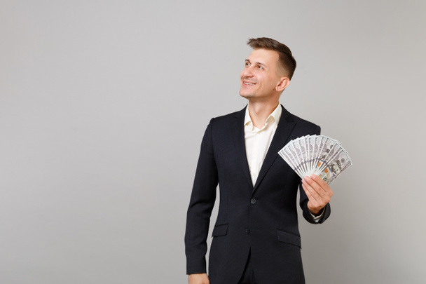 Smiling young business man in black suit looking aside, holding lots bunch of dollars banknotes, cash money isolated on grey background. Achievement career wealth business concept. Mock up copy space - Φωτογραφία, εικόνα