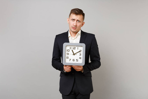 Portrait of puzzled young business man in classic black suit and shirt holding square clock isolated on grey wall background in studio. Achievement career wealth business concept. Mock up copy space - Φωτογραφία, εικόνα