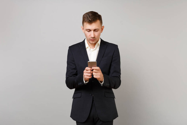 Portrait of young business man in classic black suit, shirt using mobile phone typing sms message isolated on grey background in studio. Achievement career wealth business concept. Mock up copy space - Photo, Image