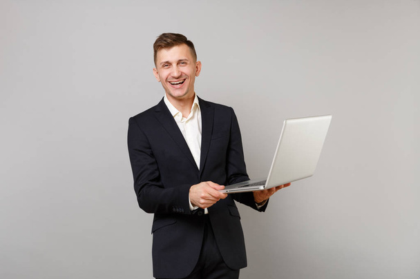 Portrait of laughing young business man in classic black suit, shirt hold laptop pc computer isolated on grey wall background in studio. Achievement career wealth business concept. Mock up copy space - Photo, image