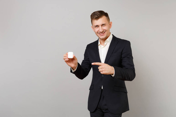 Young business man in classic black suit pointing index finger on charging case for wireless earphones isolated on grey wall background. Achievement career wealth business concept. Mock up copy space - Valokuva, kuva