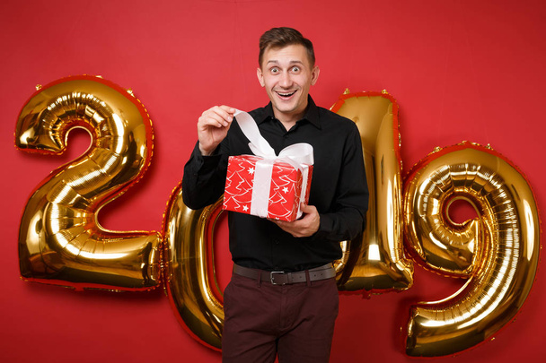 Fun man in black shirt celebrating holiday party, hold present box with gift isolated on bright red wall background, golden numbers air balloons studio portrait. Happy New Year 2019 Christmas concept - Foto, afbeelding