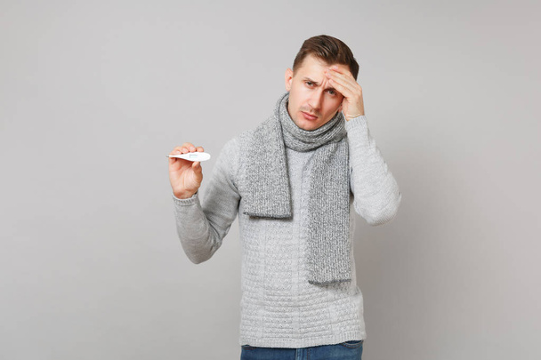 Tired young man in gray sweater, scarf holding thermometer putting hand on forehead isolated on grey background. Healthy lifestyle, ill sick disease treatment, cold season concept. Mock up copy space - Foto, imagen