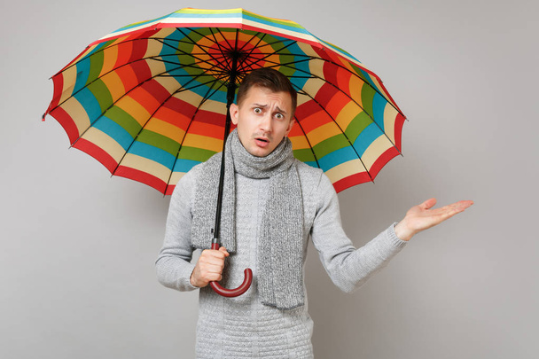 Ratloser junger Mann in grauem Pullover, Schal und buntem Regenschirm, die Hand zur Seite gestreckt auf grauem Hintergrund. Gesunder Lebensstil, Emotionen der Menschen, Konzept der kalten Jahreszeit. Attrappe Kopierraum - Foto, Bild