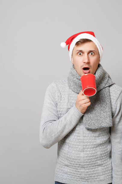 Geschokt Santa man in grijze trui sjaal KERSTMUTS rood kopje koffie of thee geïsoleerd op grijze achtergrond in de studio te houden. Gelukkig Nieuwjaar 2019 viering partij concept vakantie. Bespotten kopie databaseruimte - Foto, afbeelding