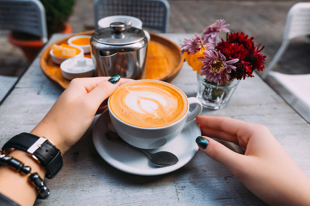 mano de niña con un reloj y una pulsera alrededor de una taza de café con espuma. Bandeja con accesorios para té y ramo de flores
 - Foto, Imagen