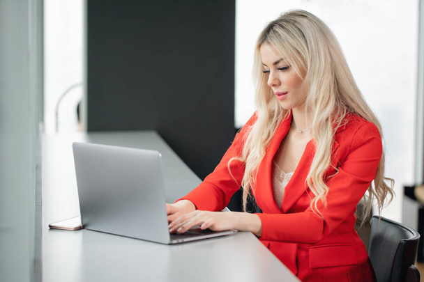 Жінка в червоному кораловому діловому костюмі. Молода блондинка з сучасним сірим ноутбуком в офісі на високому поверсі перед вікном. Дівчина п'є каву
 - Фото, зображення