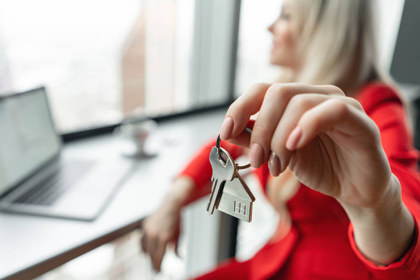 Mortgage concept. Woman in red coral business suit holding key with house shaped keychain. Modern light lobby interior. Real estate, hypothec, moving home or renting property. - Foto, imagen