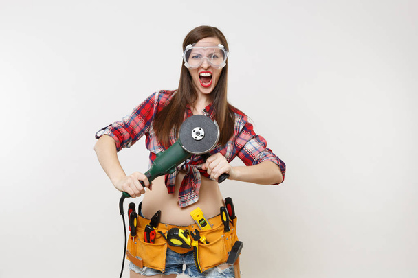 Strong young energy handyman woman in plaid top shirt, denim shorts, kit tools belt full of instruments in protective goggles hold power electric saw isolated on white background. Female in male work - Valokuva, kuva