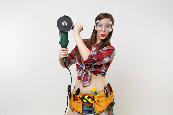 Strong young energy handyman woman in plaid top shirt, denim shorts, kit tools belt full of instruments in protective goggles hold power electric saw isolated on white background. Female in male work - Fotografie, Obrázek