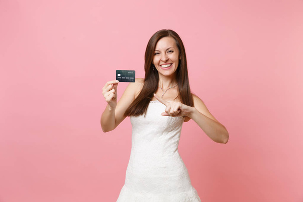 Porträt einer glücklichen Braut im weißen Brautkleid, die mit dem Zeigefinger auf die Kreditkarte zeigt, isoliert auf rosa Pastellgrund. Organisation der Hochzeitsfeier Konzept. Kopierfläche für Werbung - Foto, Bild