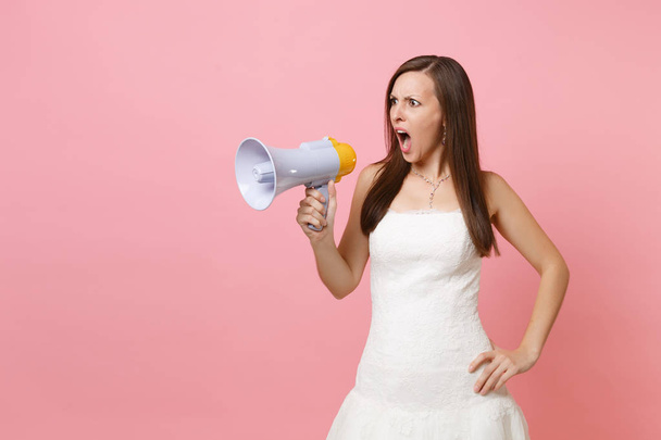Portrait of shocked bride woman in elegant white wedding dress standing and screaming in megaphone isolated on pink pastel background. Organization of wedding concept. Copy space for advertisement - Foto, Imagen