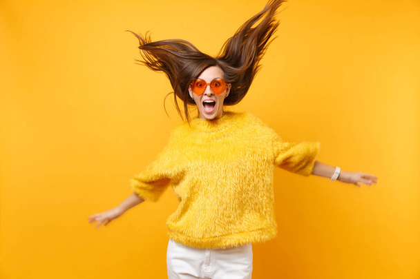 Garota excitada em suéter de pele e óculos de laranja do coração brincando no salto de estúdio com o cabelo voador isolado no fundo amarelo brilhante. Pessoas emoções sinceras, conceito de estilo de vida. Área publicitária
 - Foto, Imagem