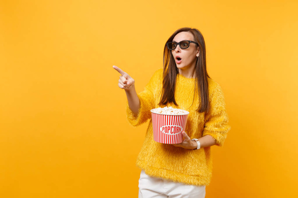 Шокированная испуганная молодая девушка в 3d imax очках указывая указательным пальцем смотреть фильм держать ведро попкорна изолированы на ярко-желтом фоне. Люди искренние эмоции в кинематографе
 - Фото, изображение