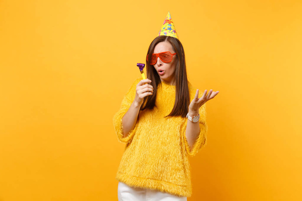 Concerned woman in orange funny glasses, birthday hat looking on playing pipe spreading hands, celebrating isolated on yellow background. People sincere emotions, lifestyle concept. Advertising area - Photo, Image
