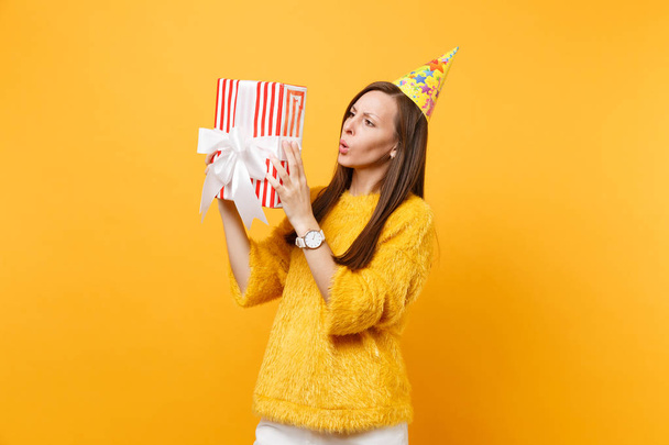 Verbaasd jonge vrouw in verjaardag feest hoed proberen te raden wat zit er in rode doos met gave presenteren vieren en te genieten van vakantie geïsoleerd op heldere gele achtergrond. Mensen oprechte emoties, lifestyle - Foto, afbeelding