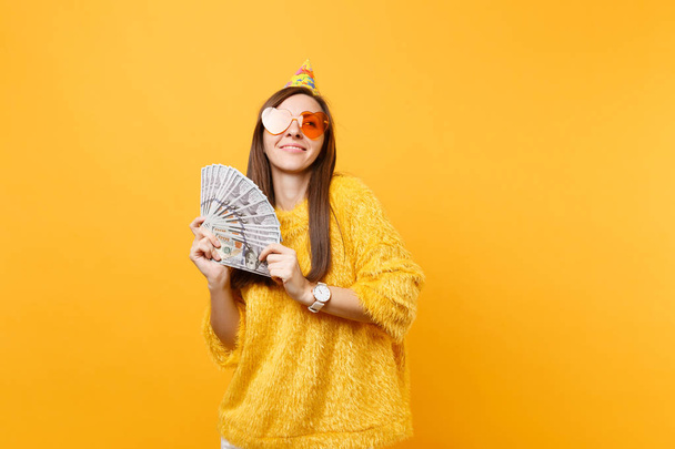 Pretty dreamy young woman in orange heart glasses birthday party hat looking up holding bundle lots of dollars cash money celebrating isolated on yellow background. People sincere emotions, lifestyle - Photo, Image