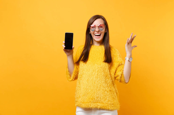 Смеющаяся молодая женщина в сердечных очках раздвигает руки и держит мобильный телефон с пустым черным пустым экраном, изолированным на ярко-желтом фоне. Люди искренние эмоции, образ жизни. Рекламная зона
 - Фото, изображение