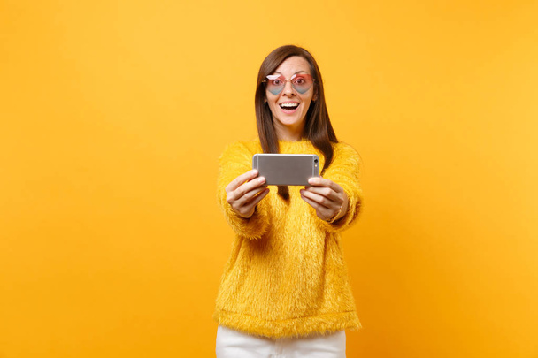 Smiling excited young woman in fur sweater, heart glasses doing taking selfie shot on mobile phone isolated on bright yellow background. People sincere emotions, lifestyle concept. Advertising area - Photo, Image