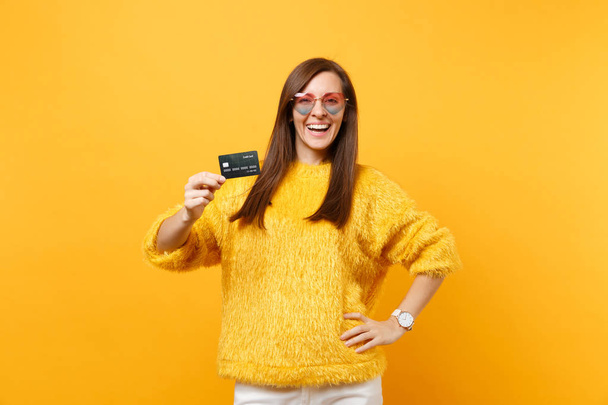 Retrato de mujer joven atractiva sonriente en suéter de piel, gafas de corazón con tarjeta de crédito aislada sobre fondo amarillo brillante. Gente emociones sinceras, concepto de estilo de vida. Área publicitaria
 - Foto, imagen