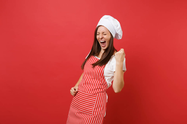 Femme au foyer cuisinière ou boulangère en tablier rayé blanc t-shirt toque chefs chapeau isolé sur fond de mur rouge. Heureuse femme expressive faisant geste gagnant, disant oui. Concept d'espace de copie simulé
 - Photo, image