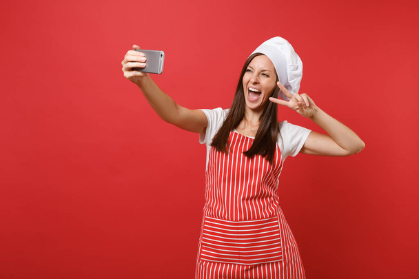 Housewife female chef cook or baker in striped apron, white t-shirt, toque chefs hat isolated on red wall background. Smiling fun woman doing selfie shot on mobile phone. Mock up copy space concept - 写真・画像